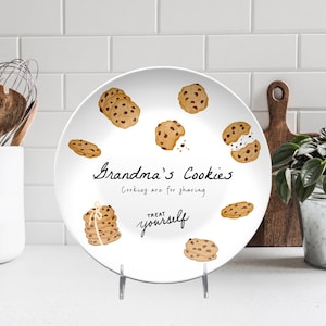 May include: White ceramic plate decorated with illustrations of chocolate chip cookies. The plate features the text "Grandma's Cookies" and "Cookies are for sharing, treat yourself" in black script. The plate is on a metal stand.