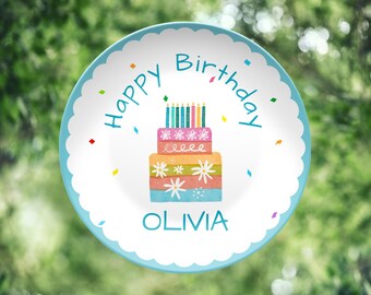 May include: A round, white plate with a scalloped edge and a light blue border. The plate features a colorful birthday cake design with candles and the words "Happy Birthday" and "Olivia" in blue.