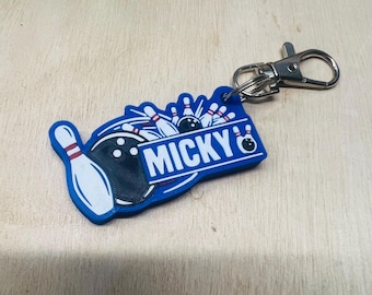 May include: A blue bowling-themed keychain with the name "MICKY" in white letters. The keychain features a bowling pin and ball design, along with a metal clasp for attachment. The background is a light wood grain.