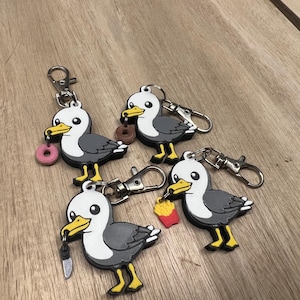 May include: Four seagull keychains made of rubber, each featuring a gray and white bird with a yellow beak and feet. The seagulls are holding different items: a pink donut, a brown donut, a small silver knife, and a red container of french fries.