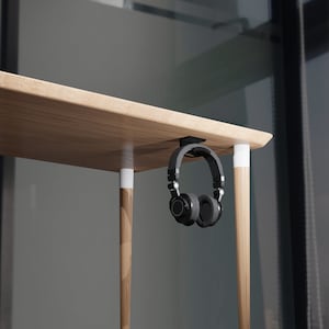 以下が含まれることがあります： 白いアクセントのある木製の机から黒いヘッドホンがぶら下がっています。机は、建物のぼやけた景色が見える窓の前にあります。