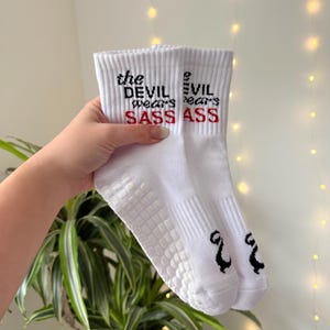 May include: White socks with the text "the DEVIL wears SASS ASS" in black and red. The socks have a textured grip on the soles and a small black graphic on the toe.