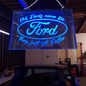 May include: A blue illuminated sign featuring the Ford logo and the text "Old Fords never Die, They just get Faster." The sign is suspended in a garage, with a classic car visible beneath it.