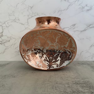 May include: A copper vase with an oval shape and a hammered finish. The vase is decorated with an intricate floral design in silver.