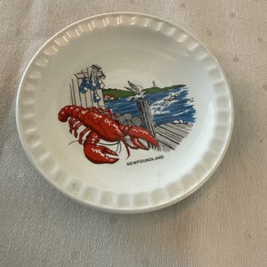 May include: A small, white ceramic dish with a scalloped edge. The dish features a colorful illustration of a red lobster, a pier, a lighthouse, and the word "NEWFOUNDLAND". The design is set against a blue and white background.