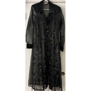 May include: Black sheer duster coat with long sleeves and a notched collar. The coat features a floral pattern with silver accents and velvet trim on the cuffs and collar. It has a button-up front and a flowing silhouette.