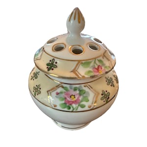 May include: A small, antique porcelain flower frog with a lid. The cream-colored jar features hand-painted pink flowers, green leaves, and gold trim. The lid has several holes for arranging flowers and a gold-tipped finial.
