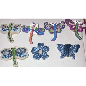 May include: A collection of ceramic decorative items featuring colorful dragonfly, butterfly, and flower designs. The dragonflies have blue, green, orange, and purple wings. The flower is blue with a cream center. The butterfly is dark blue.
