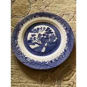 May include: A round, white and blue ceramic plate with a classic Willow pattern design. The plate features a central scene with a bridge, buildings, and trees, surrounded by a decorative border. The plate is approximately 10 inches in diameter.