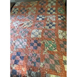 May include: A patchwork quilt featuring a grid of nine-patch blocks separated by an orange and white grid-patterned fabric. The blocks consist of various floral, striped, and checkered cotton prints in shades of blue, red, green, and white.