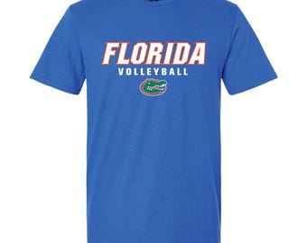 May include: A royal blue t-shirt featuring the text "FLORIDA VOLLEYBALL" in white and orange lettering. Below the text is a green and orange alligator logo. The shirt is made of a soft material.