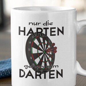 Könnte beinhalten: Weiße Keramiktasse mit einer schwarzen und grünen Dartscheibe und roten Darts. Der Text auf der Tasse lautet "nur die HARTEN gehen zum DARTEN".