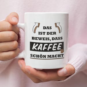 以下が含まれることがあります： 黒い文字で「Das ist der Beweis, dass Kaffee schön macht」と書かれた白いセラミック製のコーヒーカップです。