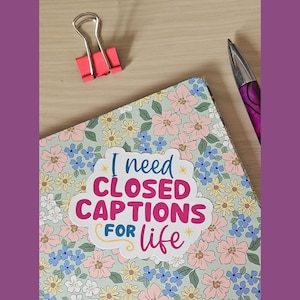 May include: A floral notebook with the text "I need CLOSED CAPTIONS FOR life." A pink binder clip and a purple pen are also visible. The notebook has a light green background with pink, blue, and yellow flowers.