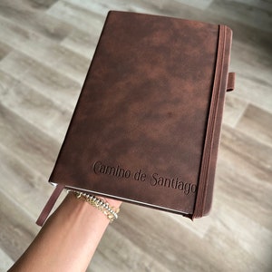 May include: A brown leather-bound journal with an elastic closure and the text "Camino de Santiago" embossed on the front cover.