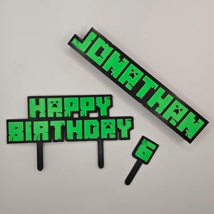 May include: Green and black birthday cake toppers with the words "HAPPY BIRTHDAY" and "JONATHAN" in a pixelated font. Includes a small number "6" topper. Ideal for a themed birthday celebration.