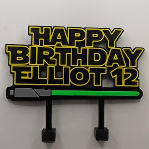 May include: A black and yellow birthday cake topper with the words "HAPPY BIRTHDAY ELLIOT 12" in a bold, futuristic font. The topper has a green bar and two black supports with square bases.