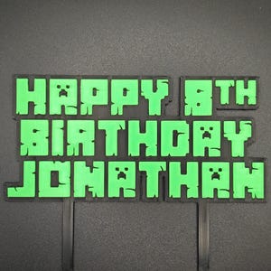 May include: A green and black cake topper with the words "HAPPY 8TH BIRTHDAY JONATHAN" in a pixelated font. The topper has two black support sticks for insertion into a cake. The text is in bright green.