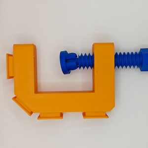 Könnte beinhalten: Orangefarbene Kunststoffklemme mit einer blauen Schraube und einem blauen Knopf. Die Klemme ist geöffnet und bereit, einen Gegenstand zu halten.