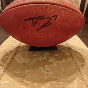 May include: A brown leather football with a black signature on it. The football is on a stand and has the NFL logo on it.