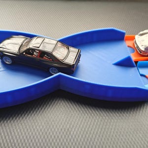 Op de afbeelding: Een blauwe plastic speelgoedautobaan met twee kleine auto's. Eén auto is zwart, de andere zilver. De baan heeft een blauw gedeelte met een X-vormig ontwerp en een oranje en blauw gedeelte.