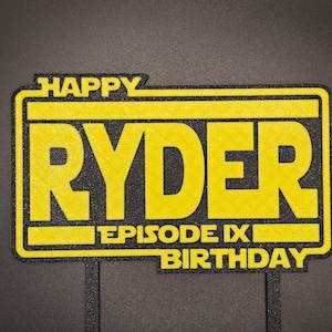 May include: A yellow and black birthday cake topper with the name "RYDER" in large yellow letters. The text "HAPPY" is above the name, and "EPISODE IX BIRTHDAY" is below. The topper has two black sticks.