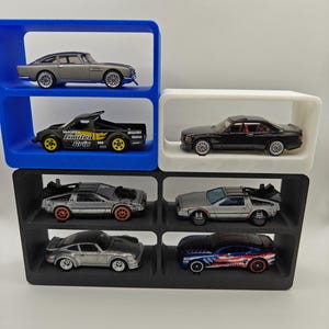 May include: A set of six black, white and blue display shelves for holding toy cars. The shelves are stacked in two rows of three. The shelves are filled with toy cars, including a silver Aston Martin, a black pickup truck with "Unlimited Grip" written on the side, a black saloon, two silver DeLorean time machines, a silver Porsche, and a blue and red Mustang.