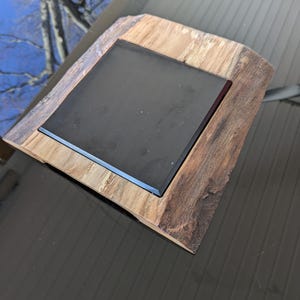 May include: A square black coaster sits on a wooden base with a natural wood grain. The base is made of a thick piece of wood with a rough edge.