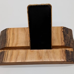 Op de afbeelding: Een houten telefoonstandaard met een natuurlijk randontwerp. De standaard is gemaakt van lichtbruin hout met een donkerbruine rand en een sleuf om een telefoon in te plaatsen. Een zwarte telefoon staat in de sleuf.