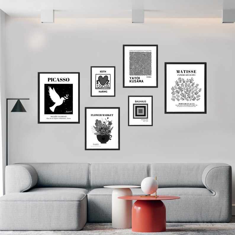 May include: A wall gallery featuring black and white framed prints of famous artists, including Picasso, Matisse, Yayoi Kusama, Keith Haring, and Bauhaus. The prints are displayed on a white wall with a gray couch and a red and white side table in the foreground.