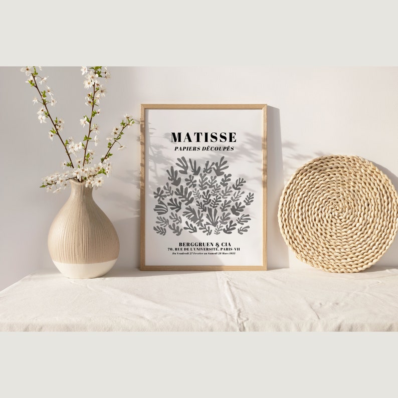 May include: A black and white poster with the text "MATISSE PAPIERS DECOUPES" and a gray abstract design of leaves. The poster is framed in a light wood frame and is sitting on a white surface.