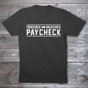 May include: A black t-shirt with the words "FORECHECK BACKCHECK PAYCHECK" printed in white block letters. The text is arranged in three lines, with the words separated by a hockey puck graphic. The shirt is made of a soft material.