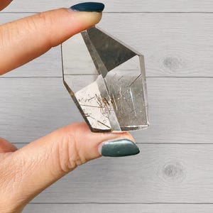 May include: A clear quartz crystal with dark inclusions and a faceted shape. The crystal is held between two fingers against a light gray wooden background. The crystal's facets reflect light, showcasing its clarity and internal features.
