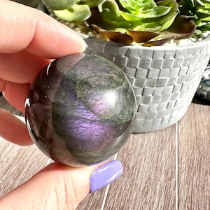 May include: A smooth, dark grey labradorite sphere with iridescent purple and green flashes. The sphere is being held in a hand.