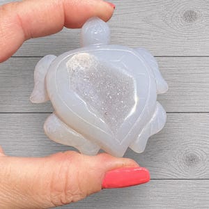 May include: A light blue, carved stone turtle figurine. The turtle has a heart-shaped design on its back, with a sparkling, crystal-filled center. The figurine is held in a person's hand against a gray wood-grain background.