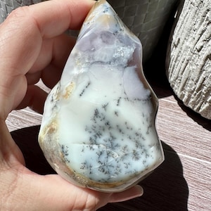 May include: A white, gray, and light purple dendritic agate freeform with a pointed top. The agate has a natural, rough texture and is a beautiful example of this type of crystal.
