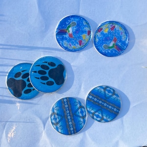 May include: Five round blue coasters with different designs. Two coasters feature a hummingbird with colorful flowers on a blue background. Two coasters feature a black bear paw print on a blue background. One coaster features a blue and white geometric pattern.