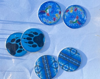 Puede incluir: Cinco posavasos redondos azules con diferentes diseños. Dos posavasos presentan un colibrí con flores coloridas sobre un fondo azul. Dos posavasos presentan una huella de pata de oso negro sobre un fondo azul. Un posavasos presenta un patrón geométrico azul y blanco.