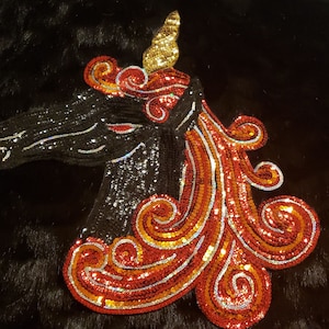 Puede incluir: Una cabeza de unicornio negra con lentejuelas, con una melena y un cuerno de lentejuelas rojas y naranjas. El unicornio está sobre un fondo negro.