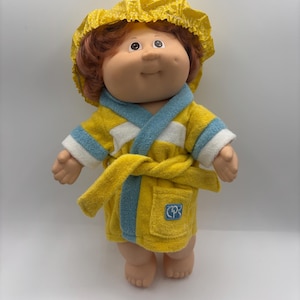 May include: A doll with reddish-brown hair wearing a yellow shower cap with white dots and a yellow terry cloth bathrobe. The robe features light blue trim, a white horizontal stripe, a tie belt, and a small pocket with a blue CP logo patch.