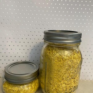 May include: Two glass jars filled with yellow nutritional yeast flakes. The jars have silver lids.