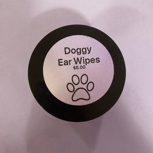 May include: A black jar with a white label that reads "Doggy Ear Wipes" with a paw print graphic. The jar is on a light purple surface.