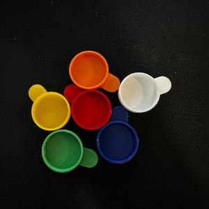 May include: A collection of colorful, small measuring scoops in yellow, red, orange, green, blue, and white. Each scoop has a small handle. The scoops are arranged on a black surface.