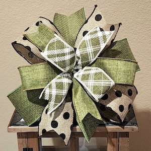 May include: A green, brown, and black plaid and polka dot bow with a burlap texture. The bow is tied with a black ribbon and is sitting on a wooden surface.