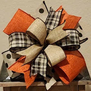 May include: A large bow made with orange, black and white plaid, and burlap ribbon. The bow is tied with a knot in the center and has a rustic look.