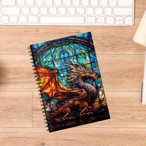 May include: A spiral bound notebook with a colorful stained glass design featuring a dragon with wings spread wide. The dragon is in shades of orange, yellow, blue, and purple.