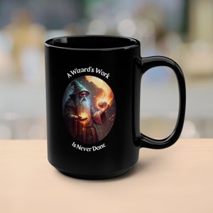 Może przedstawiać: Czarny kubek do kawy z białym wzorem przedstawiającym czarodzieja trzymającego świecącą kulę. Tekst brzmi "A Wizard's Work Is Never Done".