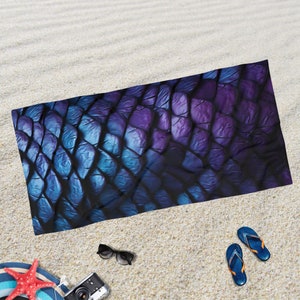 May include: A beach towel with a blue and purple iridescent scale pattern. The towel is lying on a sandy beach.