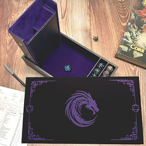 May include: A black rectangular dice box with a purple interior and a purple dragon emblem on the lid. The box is open, revealing a dice tray with a single blue die and a separate compartment containing several metallic polyhedral dice.
