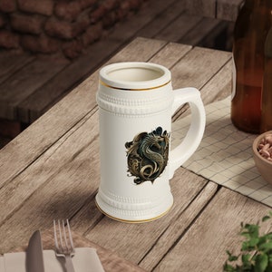 Op de afbeelding: Witte keramische bierkan met een gouden rand en een gedetailleerde illustratie van een groene en gouden draak aan de voorkant.